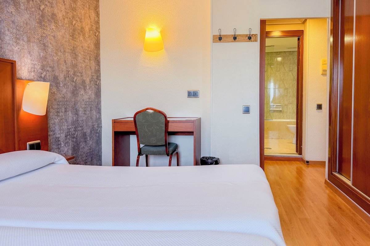 Een hotelkamer met een groot bed, een bureau met stoel, een wandlamp en een badkamer zichtbaar door een geopende deur.