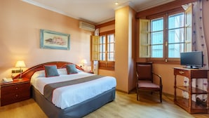 Un cuarto de hotel con una cama grande, un escritorio con televisor, una silla, una mesita de noche, una lámpara y un cuadro en la pared.