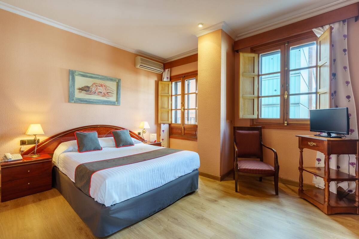 Un cuarto de hotel con una cama grande, un escritorio con televisor, una silla, una mesita de noche, una lámpara y un cuadro en la pared.