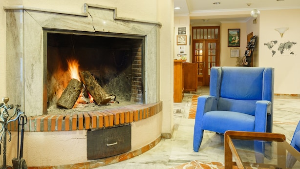 Una chimenea encendida, un sillón azul y una mesa de madera en una habitación con piso de baldosas.