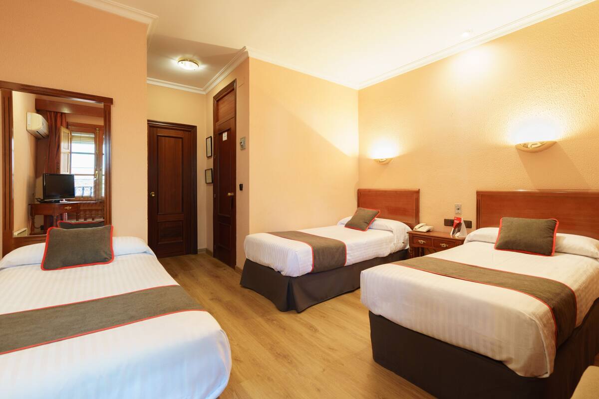 Habitación de hotel con tres camas, cada una con almohada y manta. Hay un escritorio con una silla y un televisor.