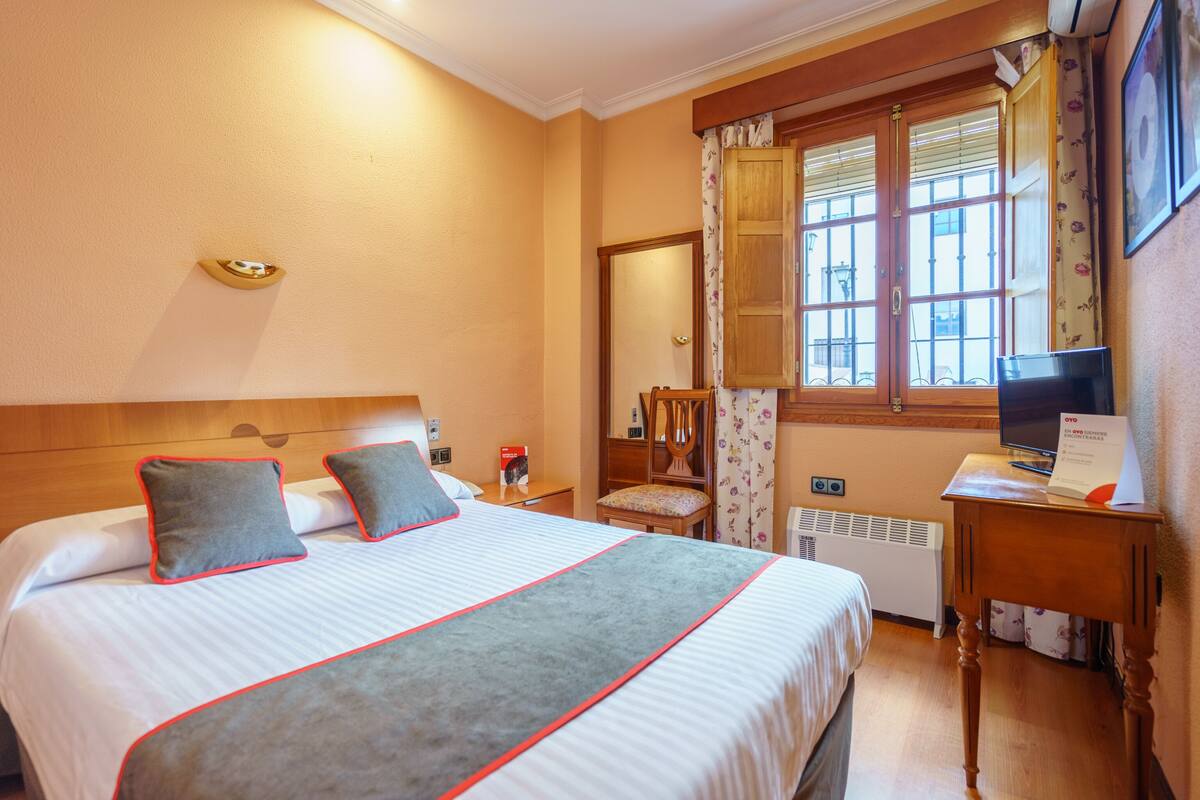 Habitación de hotel con una cama grande, cabecera de madera, un escritorio con computadora, una silla, una ventana con cortinas y un radiador.