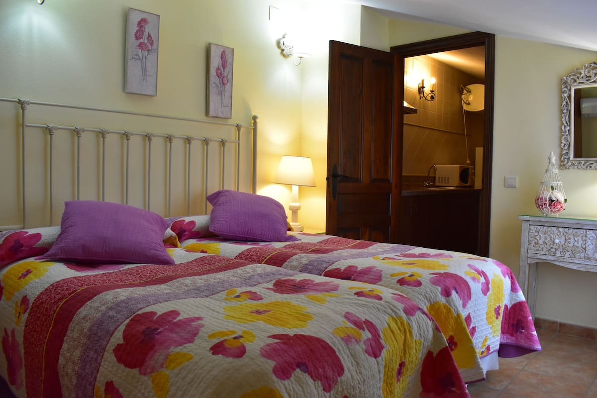 Une chambre à coucher avec deux lits, chacun recouvert d’une couvre-lit à motifs floraux et agrémenté d’oreillers violets. On y trouve une table de chevet avec une lampe, un miroir et un objet décoratif posé sur la commode.