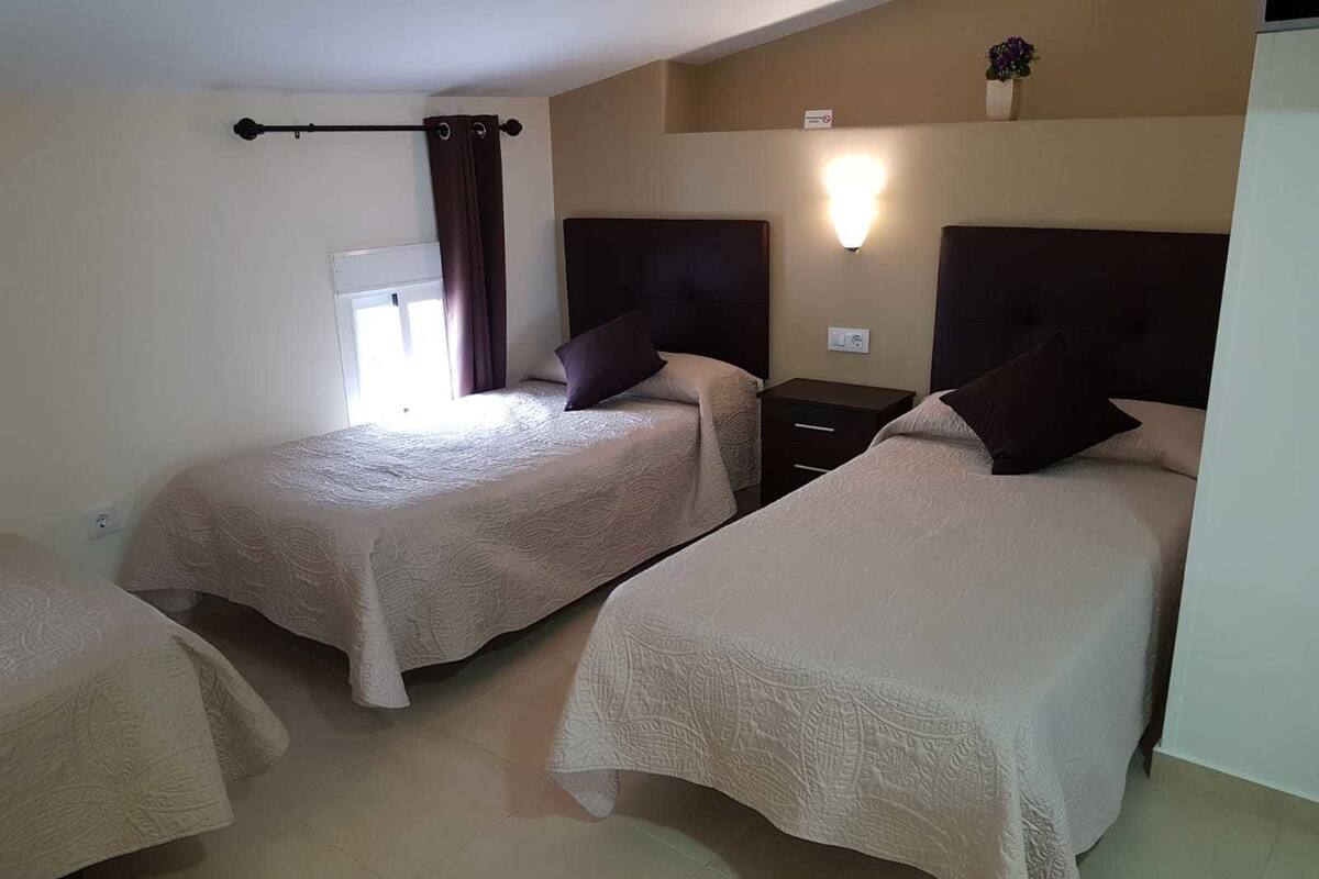 Habitación de hotel con dos camas, una mesita de noche, una lámpara de pared y una ventana con cortinas.