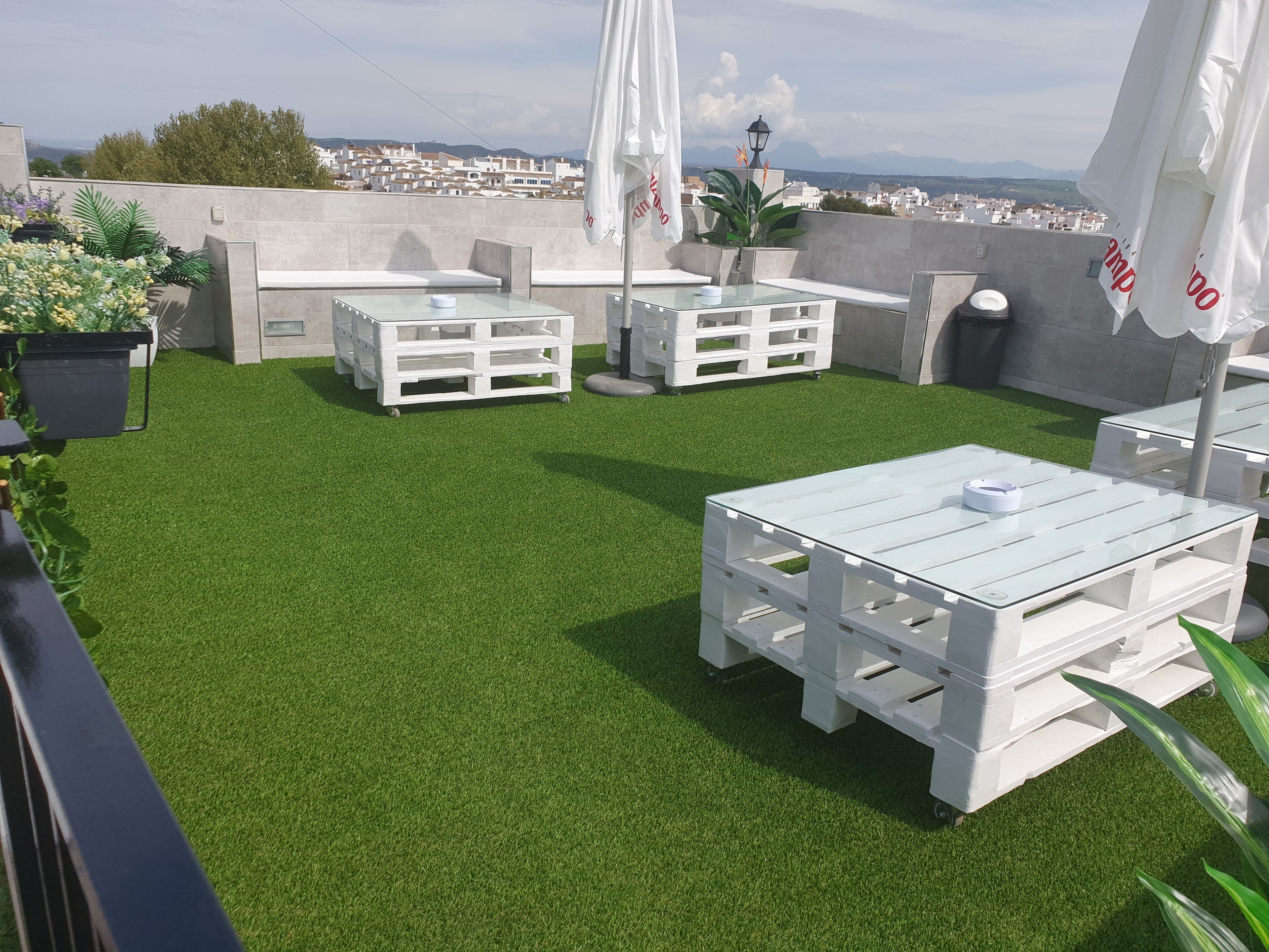 Terraza en la azotea con césped artificial, mesas y bancos blancos, y vistas de la ciudad.