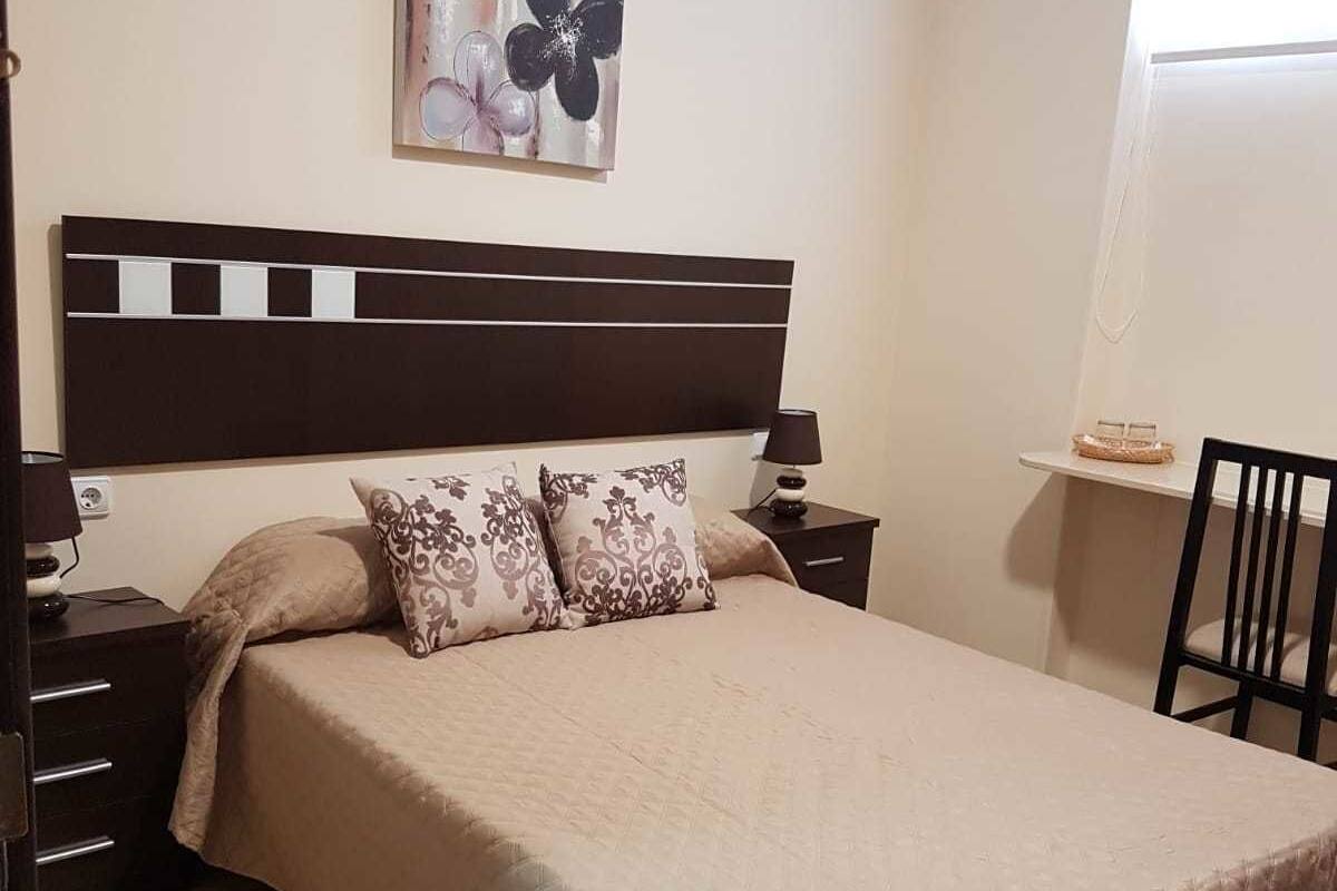 Una cama bien hecha con cubrecama acolchado, cabecero con interruptor de luz y mesita de noche con lámpara.
