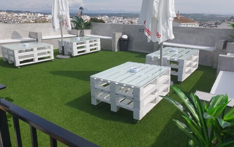 A rooftop terrace with white tables and chairs, artificial grass, and a view of a cityscape.