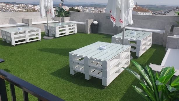 A rooftop terrace with white tables and chairs, artificial grass, and a view of a cityscape. - Hostal Málaga (Arcos de la Frontera)