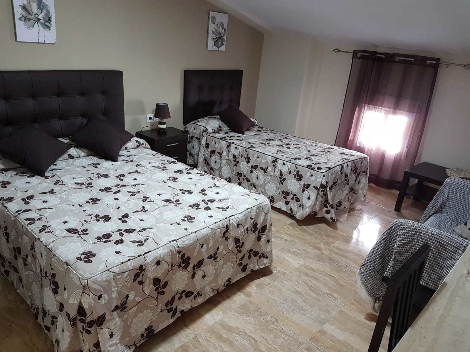 Habitació d'hotel amb dos llits, una tauleta de nit, una làmpada, una cadira i una finestra amb cortines.