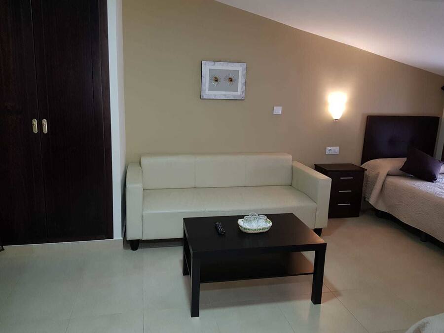 Une petite pièce bien éclairée, avec un canapé, une petite table, un lit avec une tête de lit et un luminaire fixé au mur.
