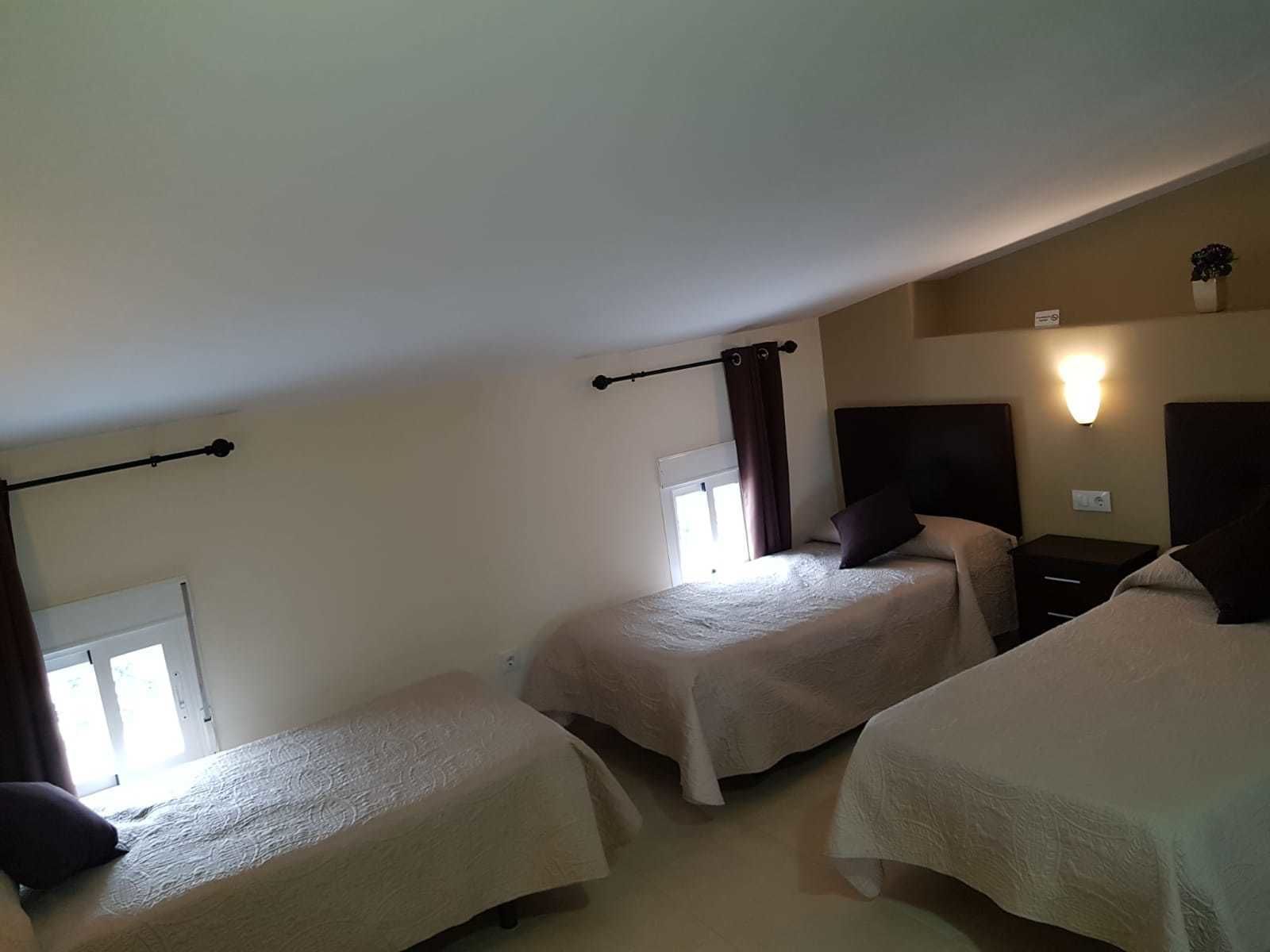 A hotel room with two single beds, a window with curtains, a nightstand, and a wall-mounted light fixture.