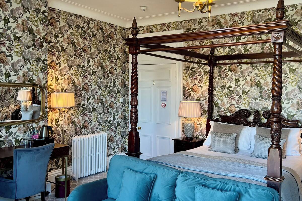 A bedroom with a four-poster bed, a blue sofa, and floral wallpaper.
