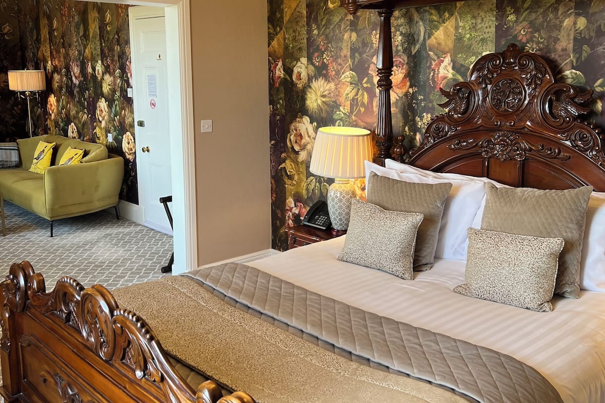 A bedroom with a large bed, ornate headboard, floral wallpaper, and a chandelier.