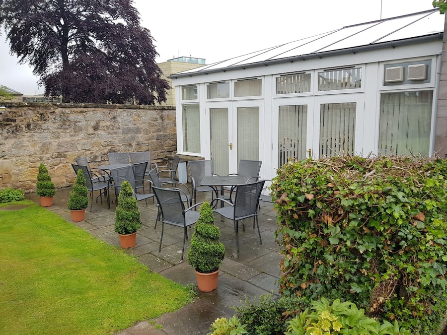 Un patio al aire libre con un muro de piedra, un edificio blanco con ventanas amplias y varias plantas en macetas.