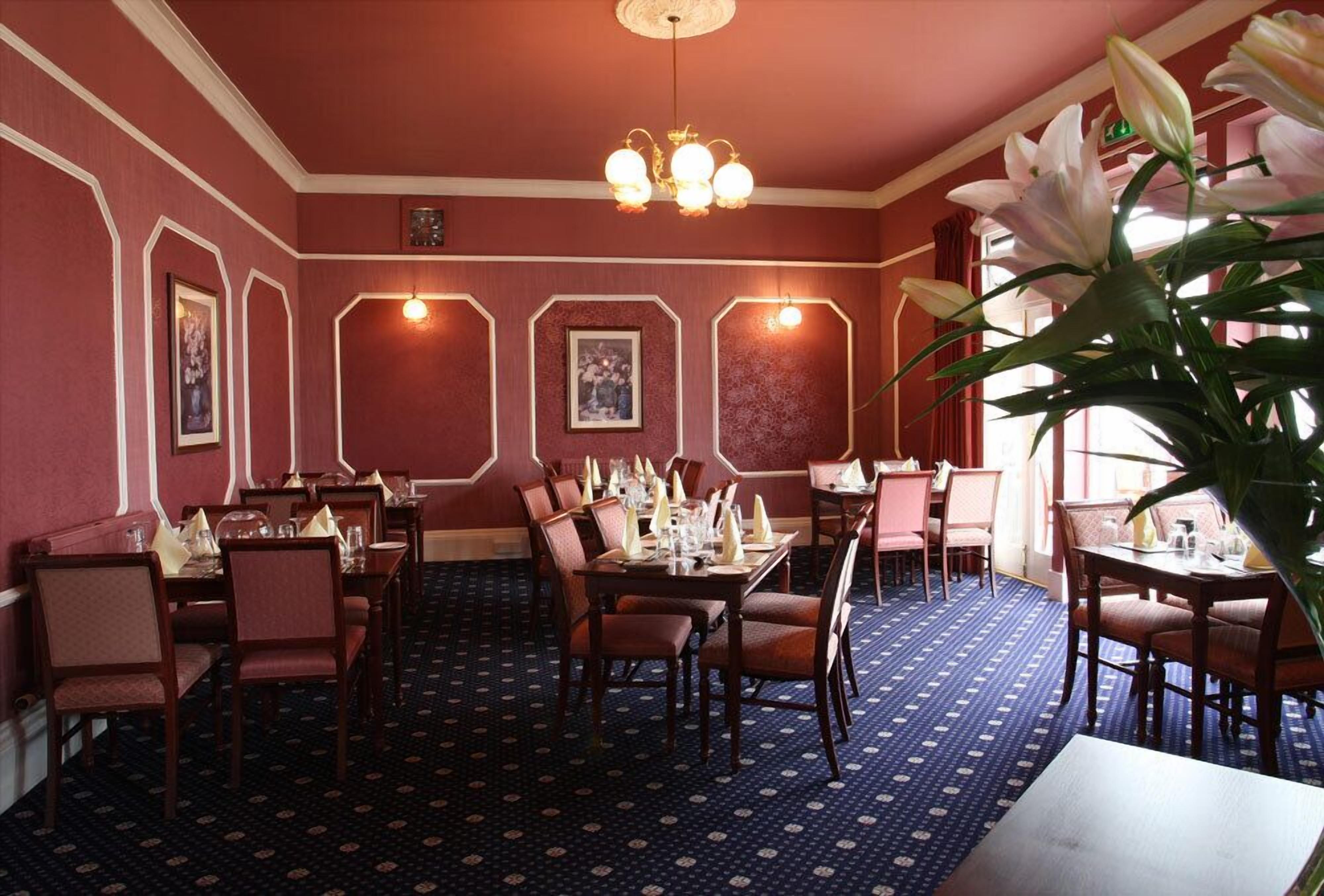 A dining area with tables set for service, framed pictures on the walls, and a patterned carpet.