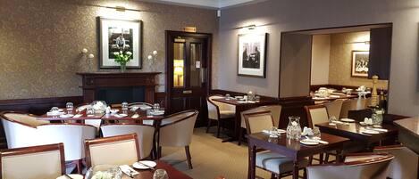 Une salle à manger avec plusieurs tables dressées, une cheminée et des tableaux encadrés aux murs.