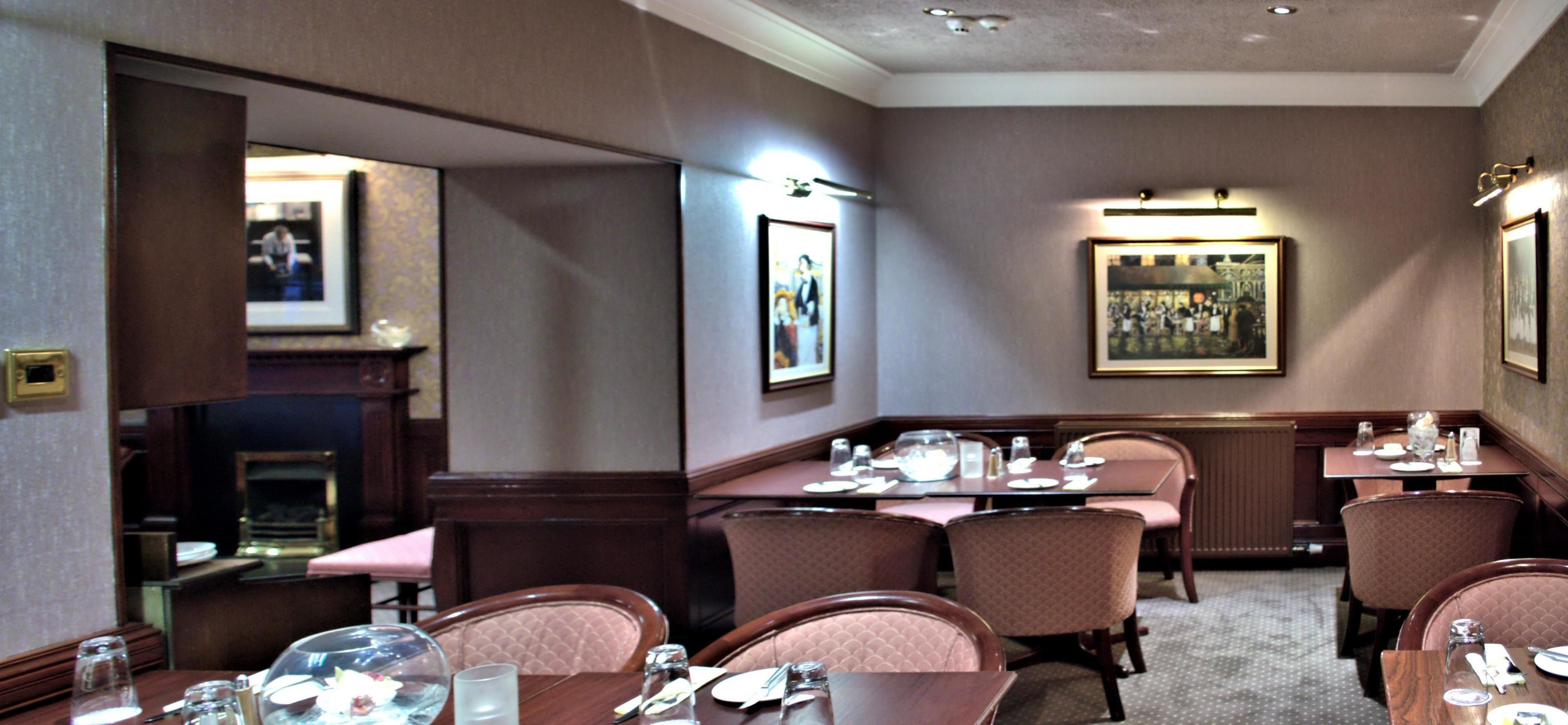 A dining area with tables set for four, each with chairs, glasses, and cutlery. There are framed pictures on the walls and a fireplace in the background.