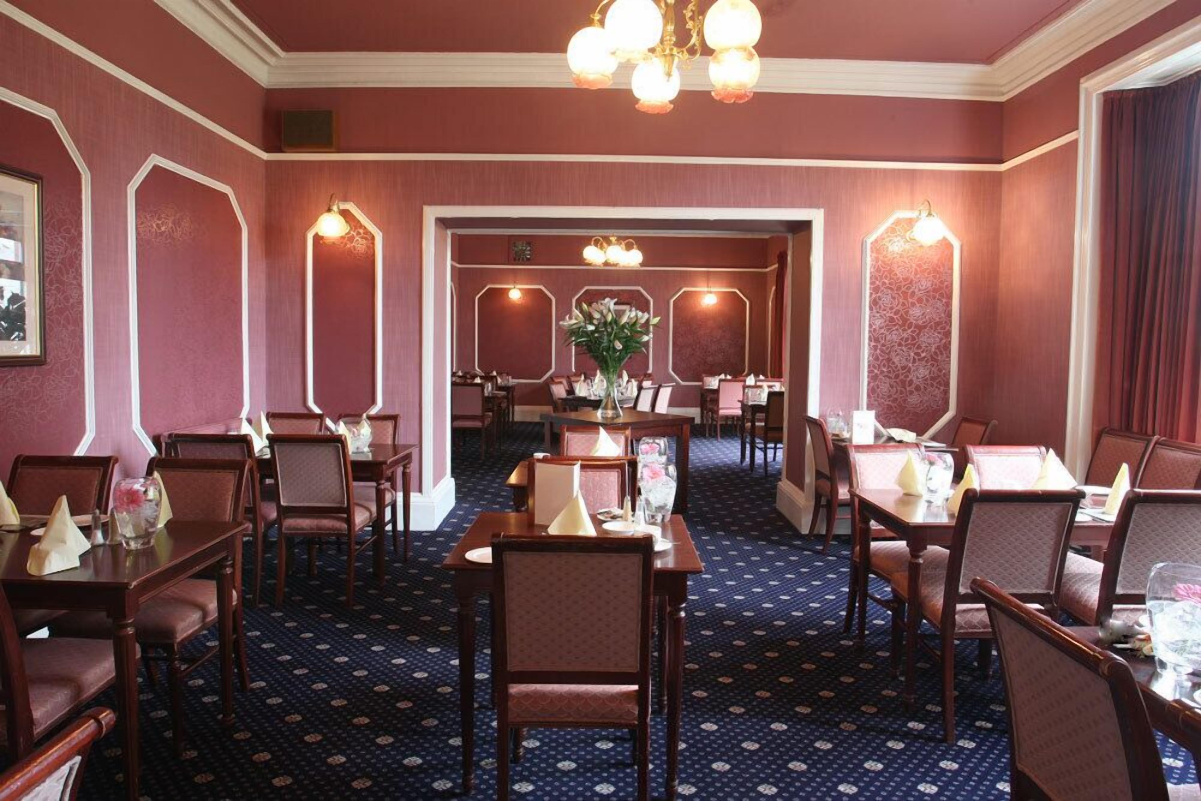 Ein Hotel-Speisesaal mit mehreren Tischen und Stühlen, einem gemusterten Teppich und dekorativen Wandleuchtern.