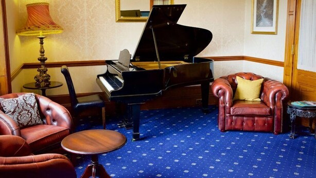 A room with a grand piano, a leather armchair, a small round table, and a lamp. - Ramnee (Forres)