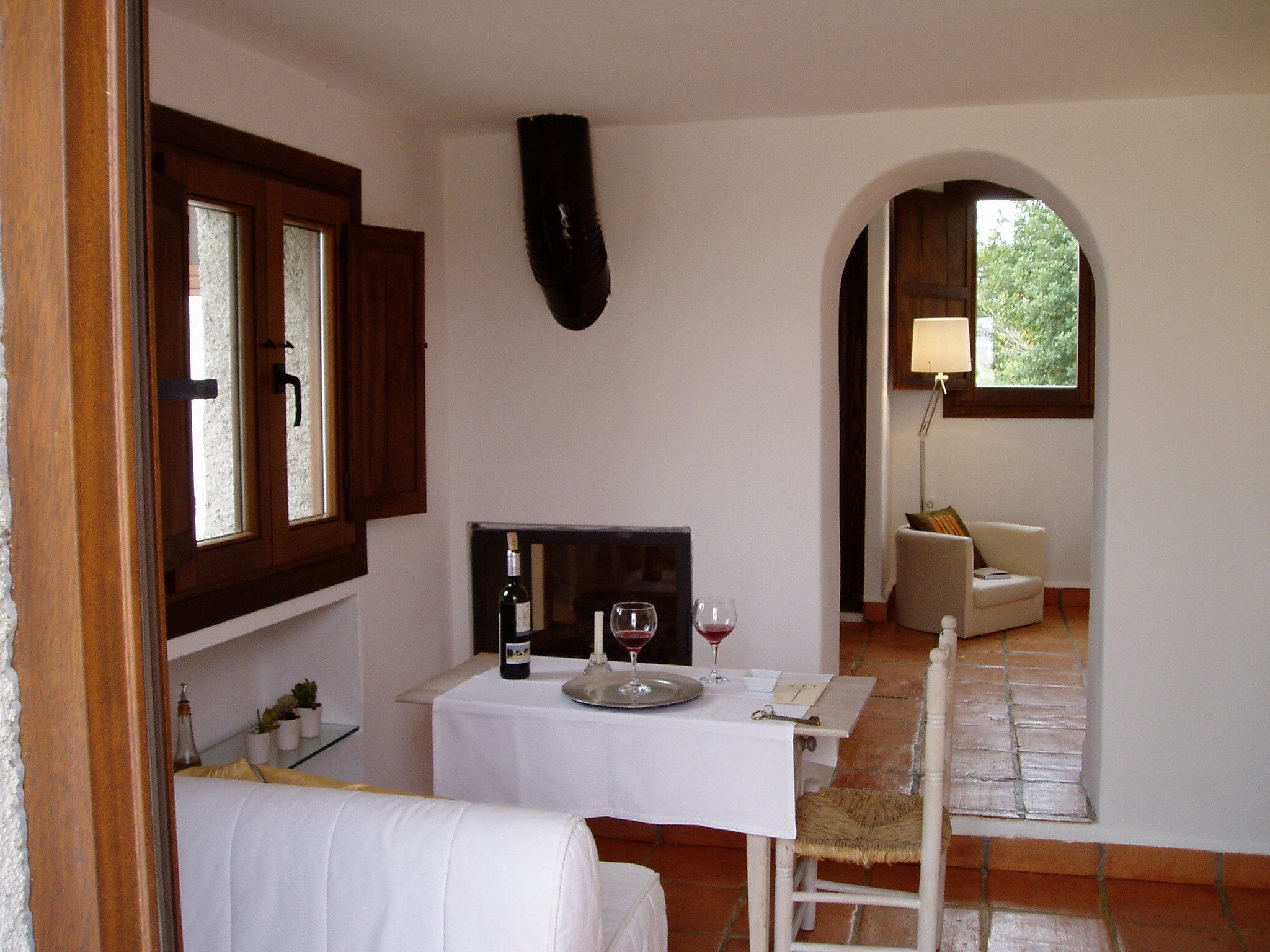 A cozy room with a dining table set for two, a bottle of wine, and a candle. There is a chair with a woven seat and a white armchair in the background.