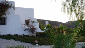 Un bâtiment blanc avec une fenêtre et une porte, entouré de verdure et bordé d’un chemin de gravier.