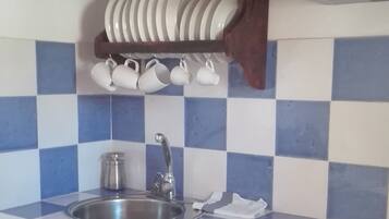 Una cocina con un fregadero, dos grifos, un estante de madera con platos y tazas, y un salpicadero de azulejos.