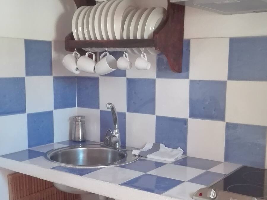 Una cocina con un fregadero, dos grifos, un estante de madera con platos y tazas, y un salpicadero de azulejos.