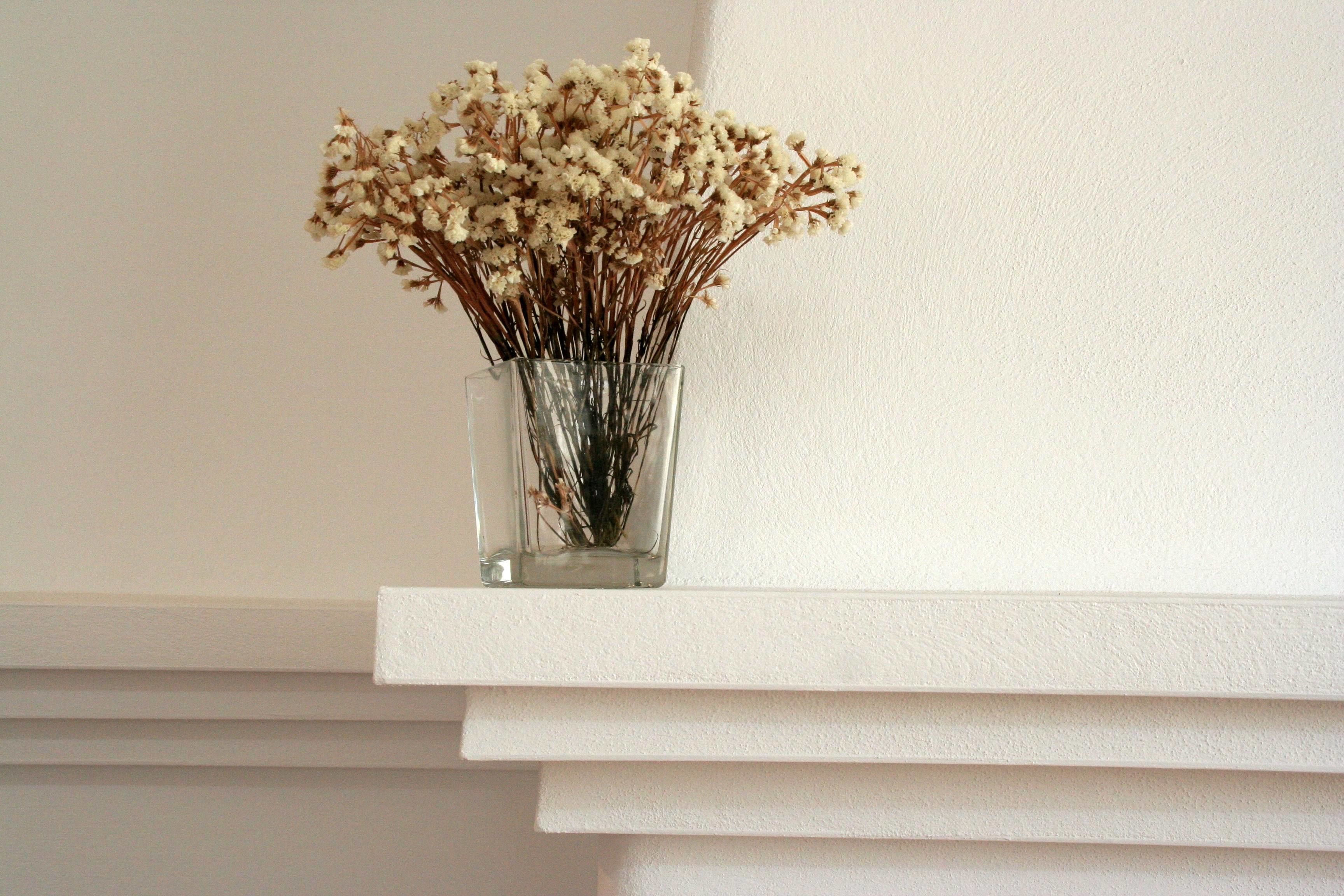 Un jarrón de vidrio transparente con flores secas sobre un estante blanco frente a una pared lisa.