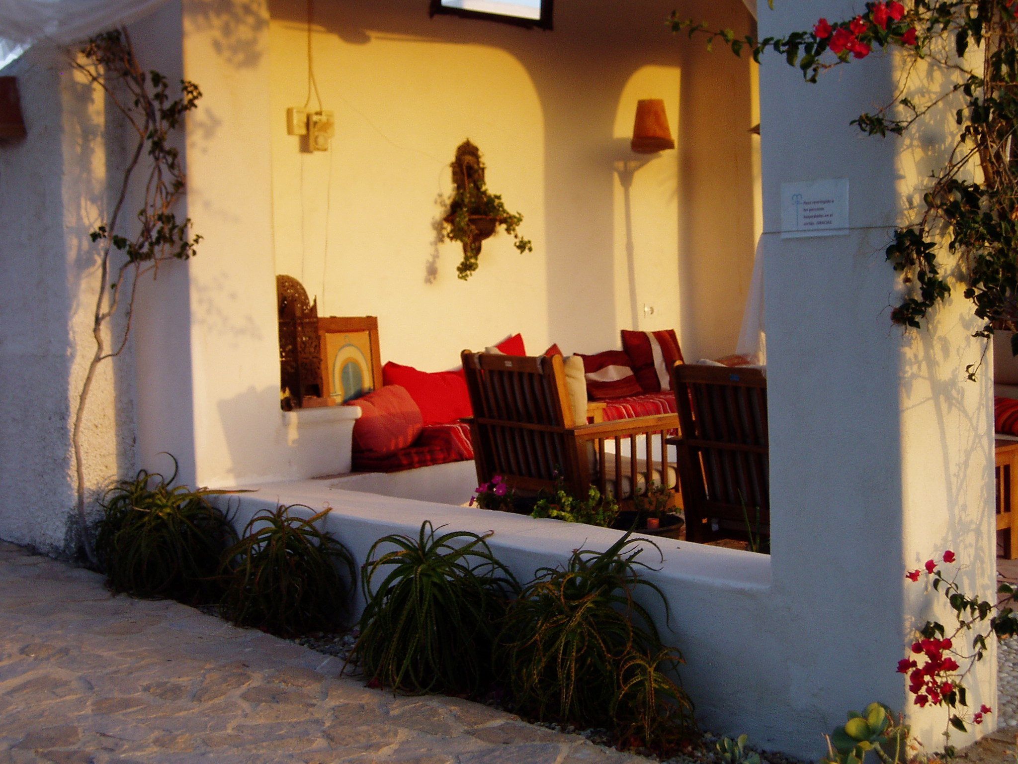 A cozy outdoor seating area with a table and chairs, surrounded by potted plants and a wall-mounted clock.