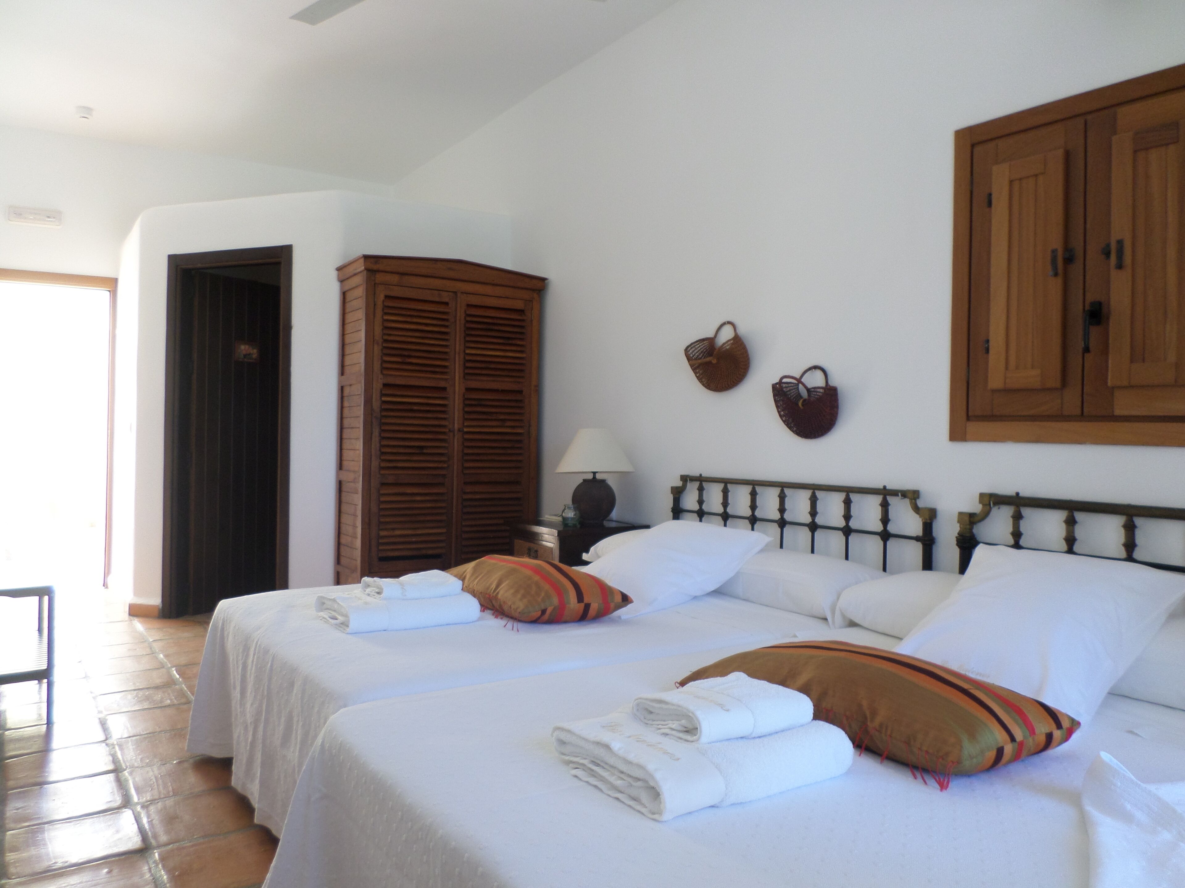 Quarto de hotel com duas camas, um guarda-roupa, uma mesa de cabeceira com luminária e uma estante fixa na parede com objetos decorativos.