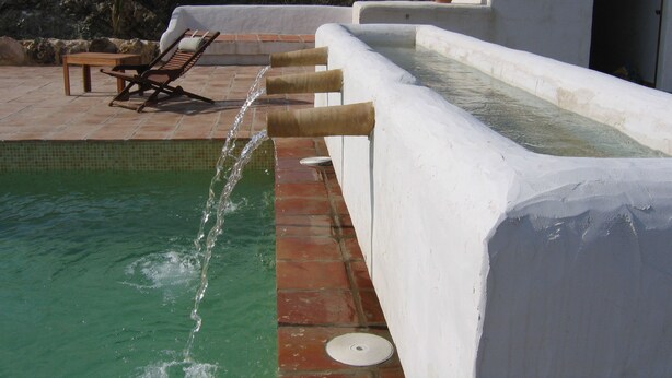 Une piscine agrémentée d’un élément décoratif aquatique, un espace de détente avec une table et des chaises, et un bâtiment blanc avec une porte ouverte.