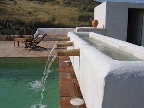 A pool with a water feature, a lounge area with a table and chairs, and a white building with an open door. - Logis Hotel Cortijo Los Malenos (Nijar)