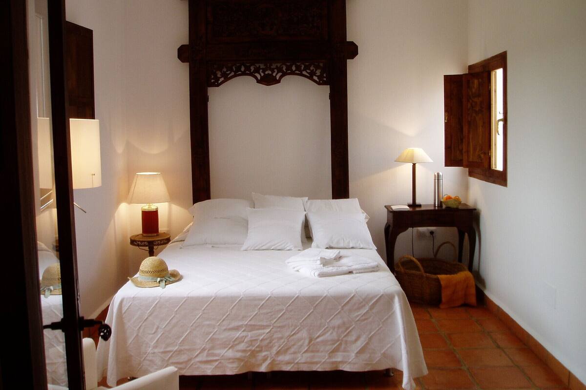 A neatly made bed with white linens, a wooden headboard, two bedside lamps, and a small table with a lamp and a basket.