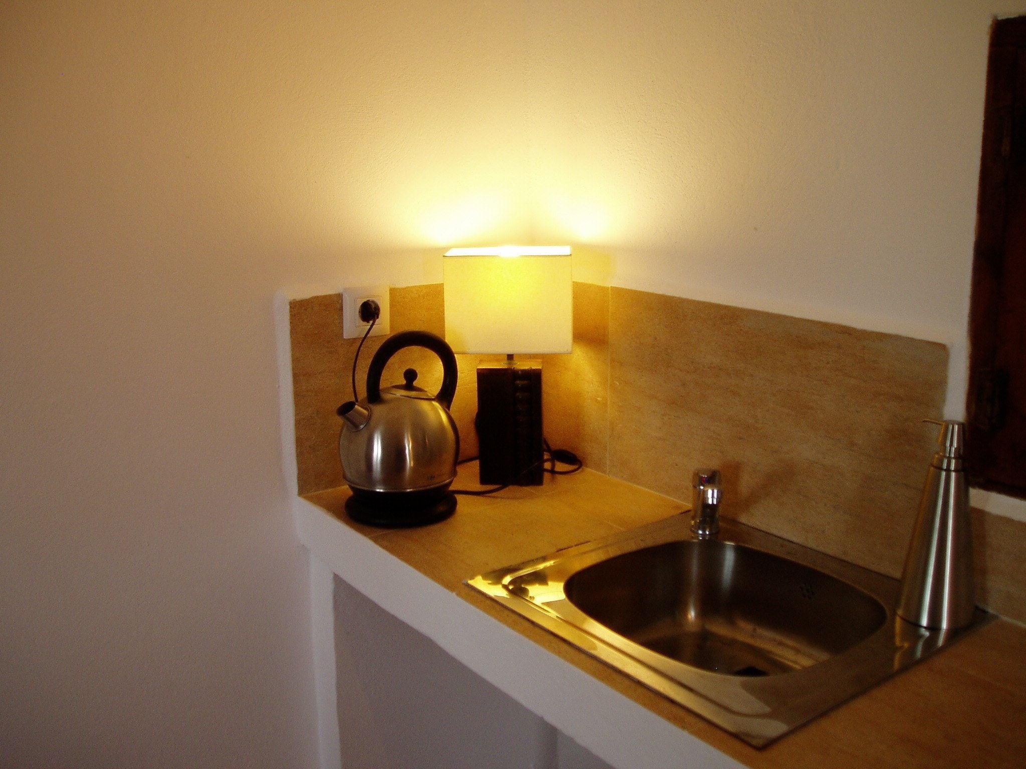 A kitchen with a stainless steel sink, a kettle, and a wall-mounted lamp.