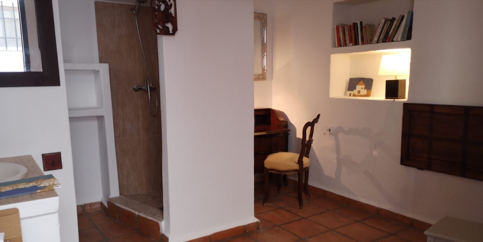 Un baño con ducha, lavabo y una silla. Hay un estante con libros y una lámpara pequeña.