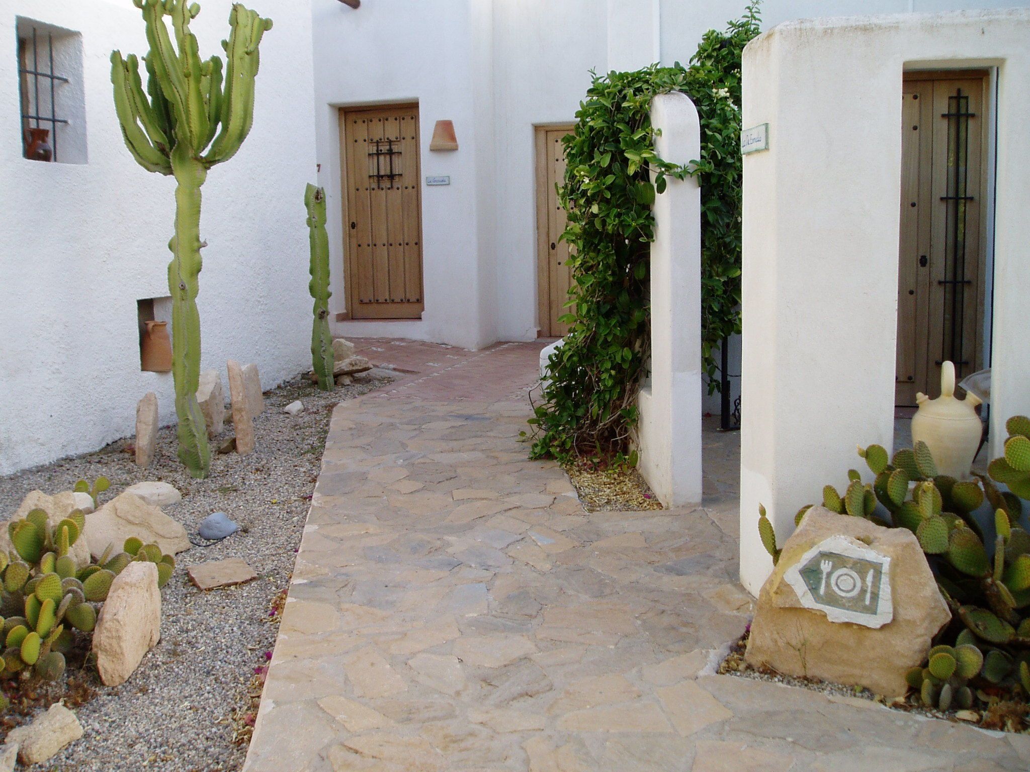 A narrow stone pathway leading to a building with white walls and wooden doors. Cacti are planted along the sides.