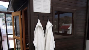 Een kleine houten hut met een glazen deur en ramen, twee witte handdoeken hangen aan haken, en een bord met tekst boven de deur.