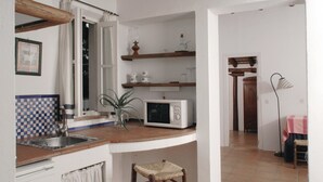Een keuken met een magnetron, een spoelbak en een klein krukje. Er is een raam met gordijnen, een plant en een plank met keukengerei.