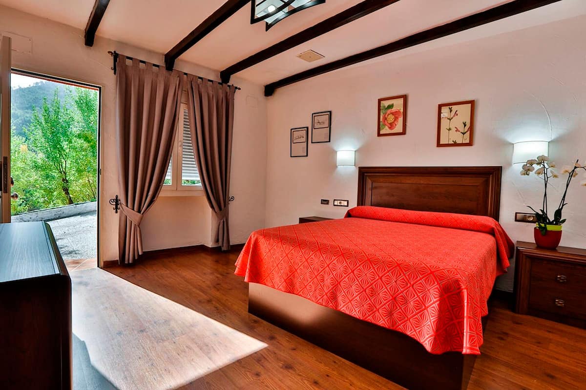 Un dormitorio con una cama grande, un escritorio, un ventanal con cortinas y vistas a zonas verdes.