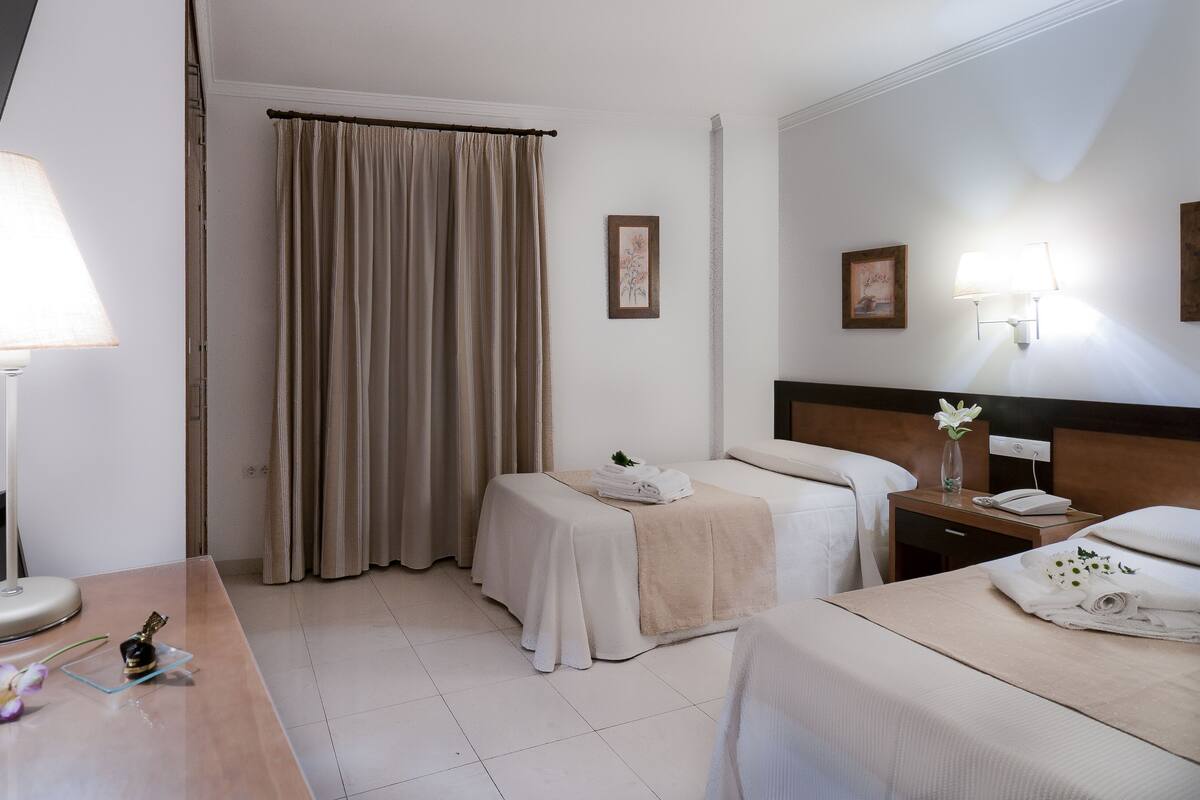 A hotel room with two beds, a desk with a lamp, a bedside table with a phone and a vase, and a large window with curtains.