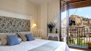 Un dormitorio con cama, una silla, una mesa con un jarrón de flores y vistas a un edificio histórico desde un balcón.