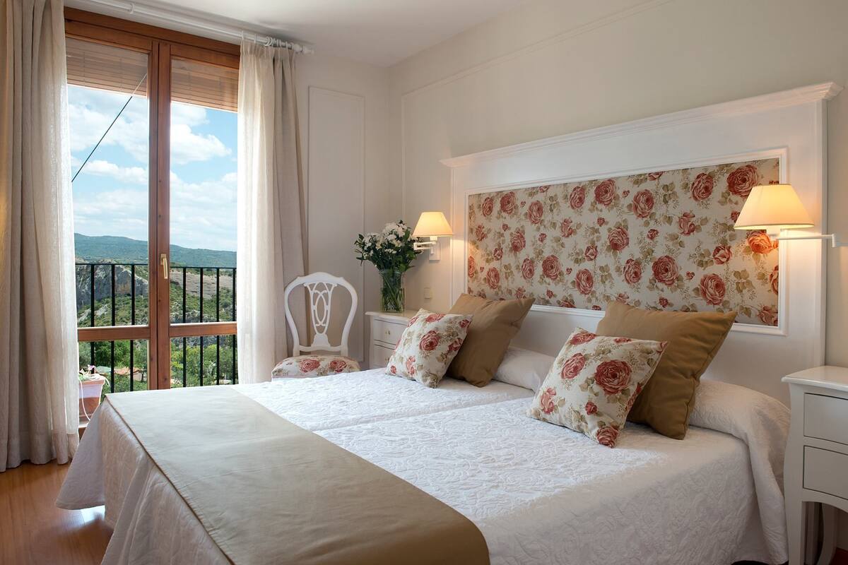 Ett hotellrum med en stor säng, två sängbord, ett litet bord, en stol, ett fönster med gardiner och en tapet med blommönster.
