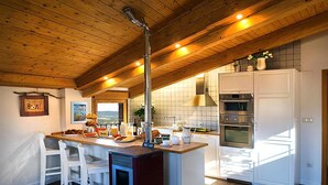 Una cocina con vigas de madera en el techo, una isla central con taburetes de bar, un horno empotrado y vistas a través del ventanal.
