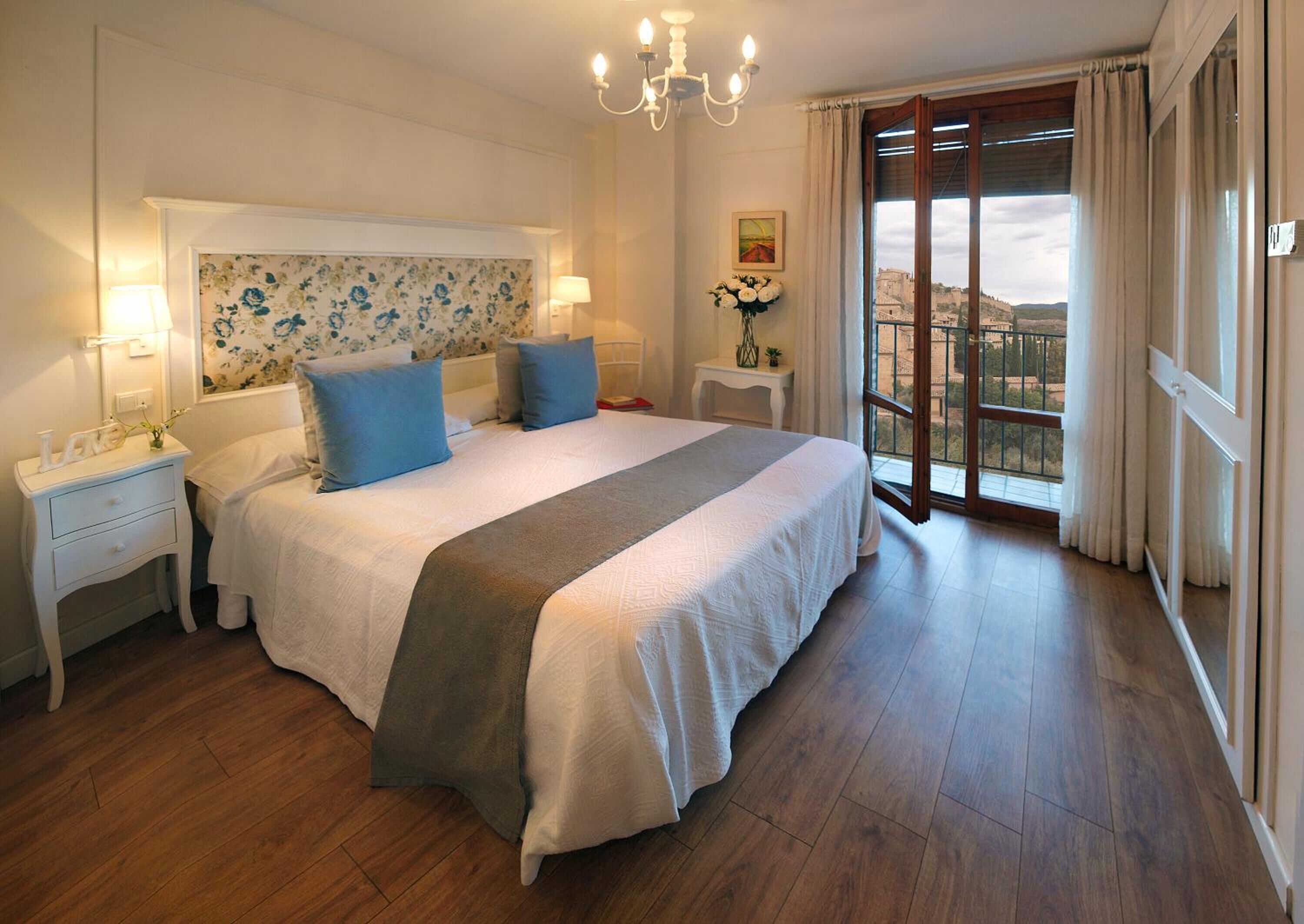 A bedroom with a large bed, two bedside tables, a chandelier, a window with curtains, and a view of a building outside.