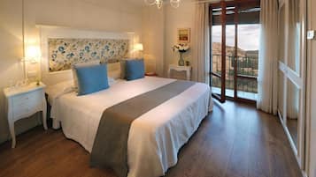 A bedroom with a large bed, two bedside tables, a chandelier, a window with curtains, and a view of a building outside.