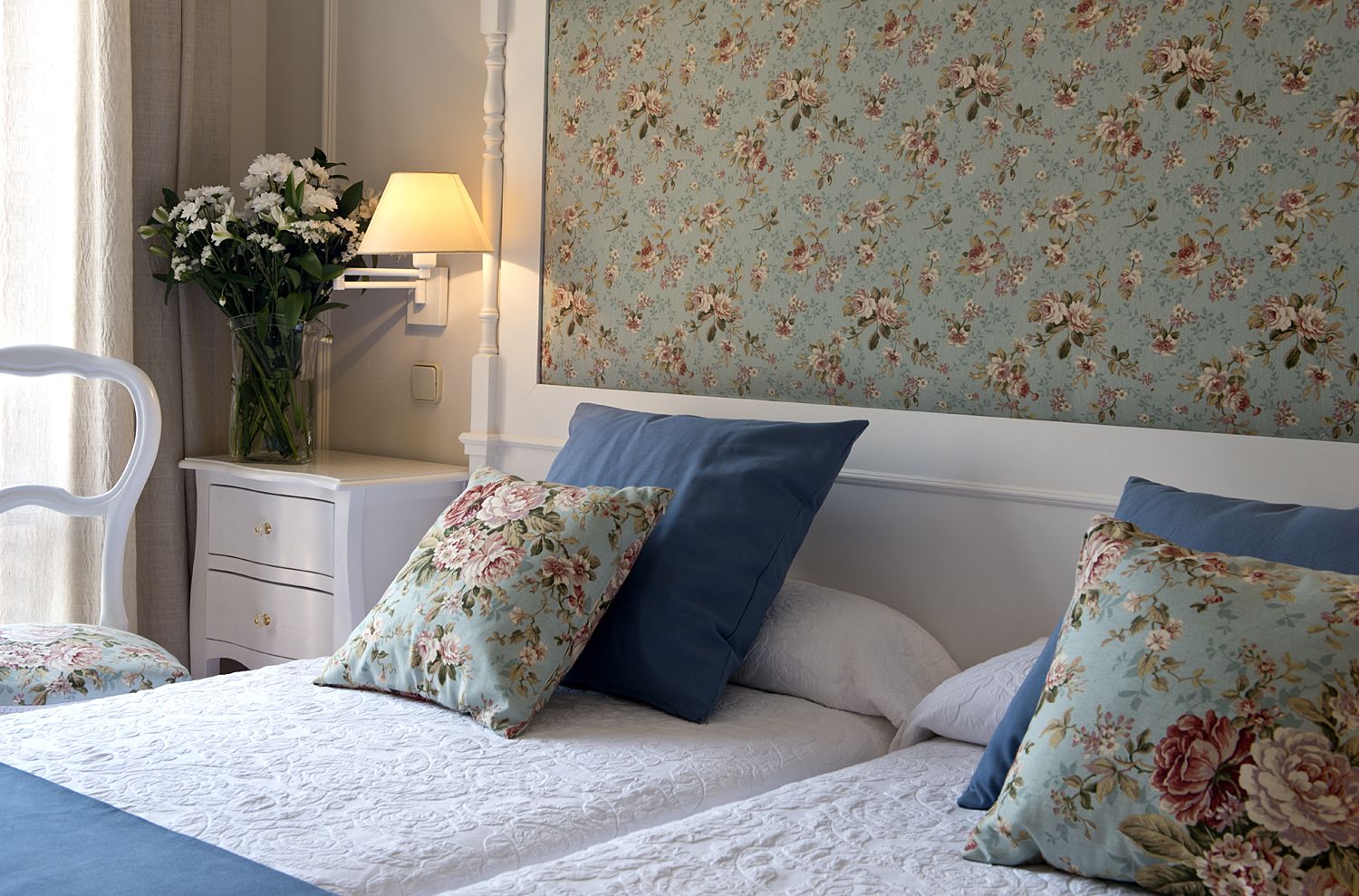 Un dormitorio con un cabecero estampado con motivos florales, una mesita de noche con lámpara y un florero con flores.