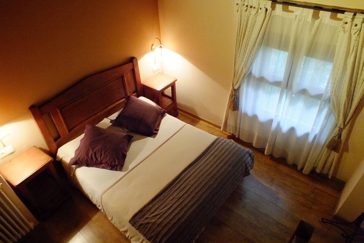 A neatly made bed with a wooden headboard, two pillows, and a blanket. There is a nightstand with a lamp, a window with curtains, and a chair in the corner.