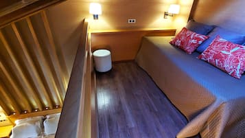 Interni di un camper compatto con pavimento in legno, una cuccetta con cuscini a motivi rossi e blu e due luci a parete.