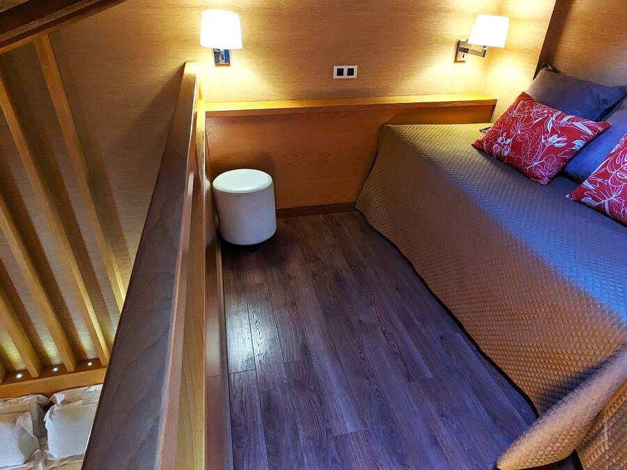 Interni di un camper compatto con pavimento in legno, una cuccetta con cuscini a motivi rossi e blu e due luci a parete.