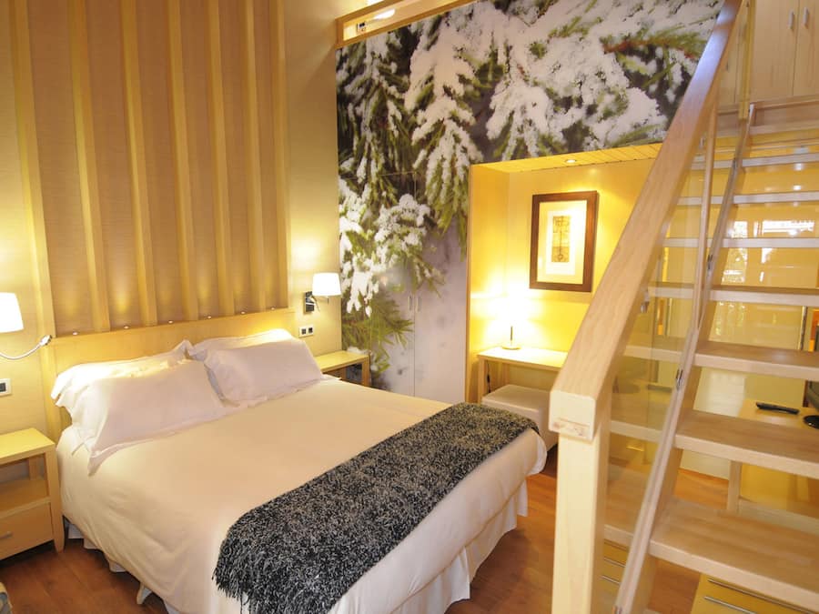 Una habitación de hotel con una cama, mesitas de noche, una lámpara, un cuadro y una escalera de madera.